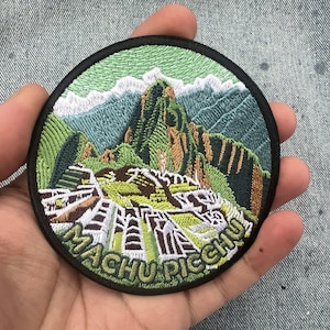 May include: A circular embroidered patch featuring a detailed landscape of Machu Picchu with green mountains, stone ruins, and white snow-capped peaks. The text "MACHU PICCHU" is stitched in light green letters along the bottom edge with a black border.