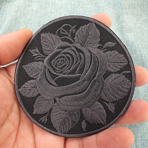 May include: A circular embroidered patch featuring a dark grey rose design with detailed petals and leaves centred on a black background. The embroidery uses tonal thread to create depth and texture within the floral motif, finished with a stitched border.