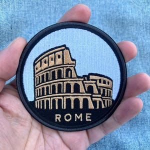 Könnte beinhalten: Ein runder, bestickter Aufnäher mit einer goldfarbenen Darstellung des Kolosseums vor einem hellblauen Hintergrund. Der Aufnäher hat einen dicken schwarzen Rand und das Wort ROME ist in goldenen Großbuchstaben unten mittig eingestickt.