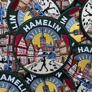 Peut inclure: Un écusson brodé circulaire représentant une scène de Hamelin avec des maisons à colombages, une église centrale et un pont. La silhouette d'un joueur de flûte mène un groupe de rats au premier plan. Le mot HAMELIN est brodé en blanc.