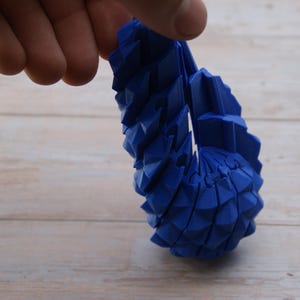 Könnte beinhalten: Ein flexibles, bewegliches blaues Objekt mit einer segmentierten, geometrischen, schuppenartigen Textur. Das Stück besteht aus mehreren miteinander verbundenen, sich wiederholenden dreieckigen Einheiten, die es biegen und krümmen.