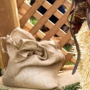 May include: A tan burlap sack with a gathered top opening, positioned next to a brown leather saddle and a pile of light yellow straw. The items are placed in front of a light-colored wooden lattice fence structure on a patch of green grass.