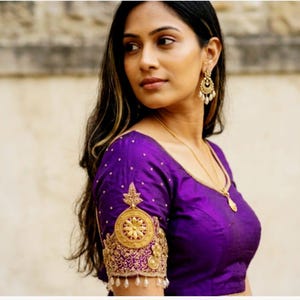 May include: A vibrant purple blouse featuring intricate gold embroidery on the sleeves, accented with small white beads and dangling pearl-like drops. The garment includes a round neckline and is paired with gold drop earrings and a gold necklace.