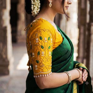Puede incluir: Una blusa de color amarillo mostaza con mangas cortas, bordados florales blancos y detalles de hojas verdes, combinada con un sari verde oscuro. La blusa tiene un dobladillo festoneado con cuentas blancas colgantes y brazaletes dorados.