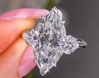 May include: A silver-toned ring featuring a large marquise-cut center stone held by prongs, flanked by two smaller round-cut stones on either side. The band is polished metal, and the stones are clear and faceted, reflecting light from multiple angles.
