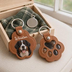 Puede incluir: Dos llaveros de cuero color canela con forma de pata y anillas de metal plateado. Uno muestra el retrato impreso de un perro de montaña de Berna y el nombre Winston en letra blanca. El otro tiene una ventana transparente con pelo.