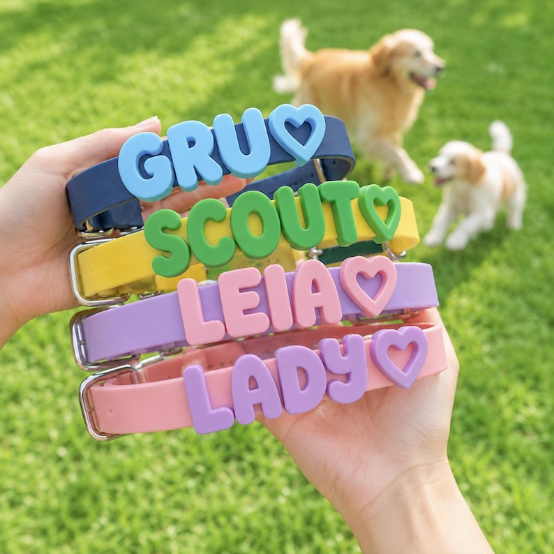 May include: A stack of four silicone dog collars in navy blue, yellow, lavender, and light pink. Each collar features raised, block-letter names reading GRU, SCOUT, LEIA, and LADY, with a matching heart-shaped charm attached to the end of each name.
