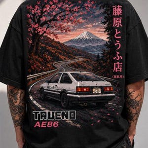 May include: A black t-shirt featuring a graphic print of a white Toyota AE86 Trueno car on a winding road, with a mountain and cherry blossom tree in the background. The text reads "TRUENO AE86" and Japanese characters are printed on the right side.