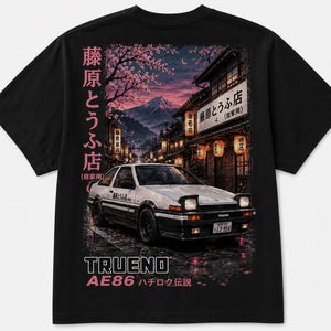 May include: A black t-shirt featuring a graphic print of a white and black AE86 Trueno car parked on a wet street at night. The background shows a Japanese street scene with cherry blossoms, lanterns, and a mountain. Text includes "TRUENO" and Japanese characters.
