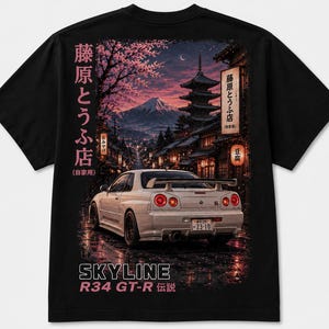 May include: A black t-shirt featuring a graphic print of a white Nissan Skyline R34 GT-R parked on a wet street. The background shows a Japanese pagoda, Mount Fuji, and cherry blossoms under a night sky. Japanese text and the words SKYLINE R34 GT-R are visible.