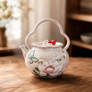 Può includere: Una teiera in ceramica rosa chiaro con un manico alto e arcuato e un coperchio con un piccolo dettaglio a perla rossa. Il corpo è decorato con motivi floreali in rilievo bianchi e rosa e una farfalla verde, rifinito con dettagli dorati.