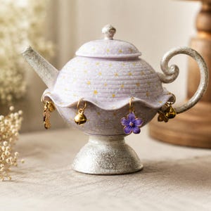 Può includere: Una teiera in ceramica color lavanda con un motivo a margherite bianche, beccuccio, manico e base dalla texture argentata. La teiera presenta un bordo arricciato decorato con diversi piccoli ciondoli pendenti, tra cui campanellini dorati.