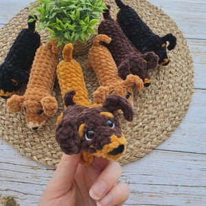May include: A collection of six handmade crochet dachshund plush toys in black and various shades of brown. Each stuffed animal features a soft, textured yarn finish, floppy ears, and small black eyes. The toys are arranged in a circular pattern.