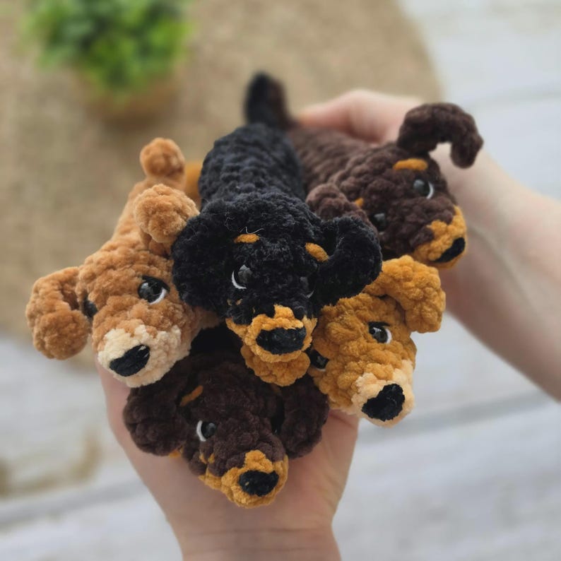 May include: A collection of five small, plush, crocheted dachshund toys made from soft, fuzzy yarn. The dogs feature various color combinations including solid black, dark brown, and light tan, each with black noses and dark eyes.