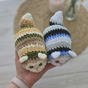 May include: Two small, plush, crocheted animal figures with soft, fuzzy yarn. One figure features horizontal stripes of cream, tan, and dark green. The other figure has stripes of white, black, gray, and blue. Both have small ears and stitched faces.