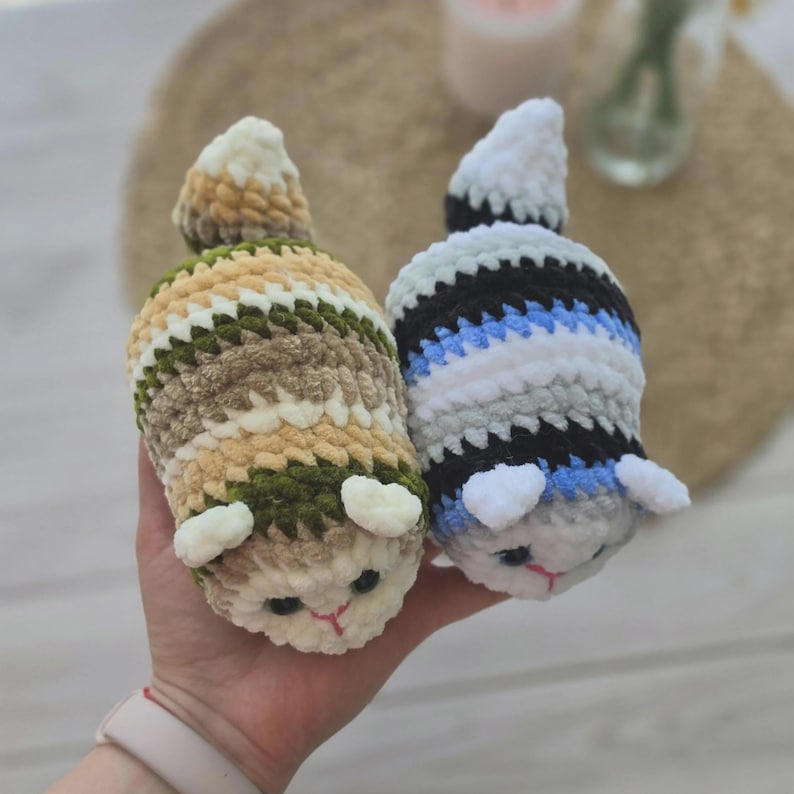 May include: Two small, plush, crocheted animal figures with soft, fuzzy yarn. One figure features horizontal stripes of cream, tan, and dark green. The other figure has stripes of white, black, gray, and blue. Both have small ears and stitched faces.