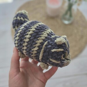 May include: A small, handmade crochet plush toy shaped like a cat. The figure is crafted from soft, chunky yarn in alternating stripes of dark charcoal gray and light beige. It features a rounded body, a short tail, small ears, and dark eyes.
