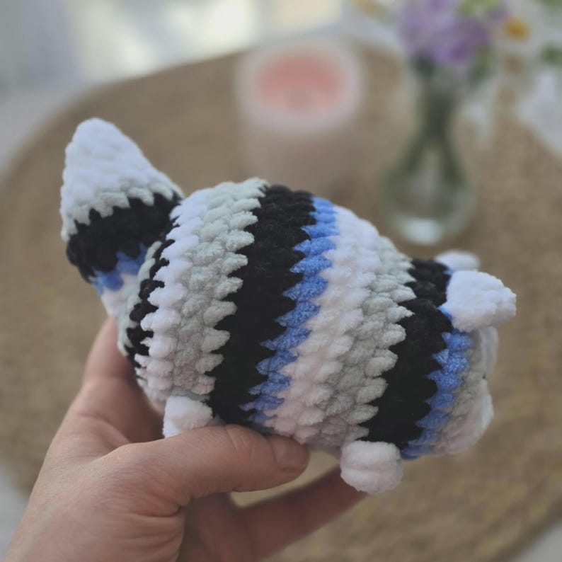 May include: A small, plush, crocheted animal figure featuring a striped pattern of white, light gray, black, and light blue yarn. The soft, textured toy has a rounded body, a pointed tail, and small white appendages extending from the sides.