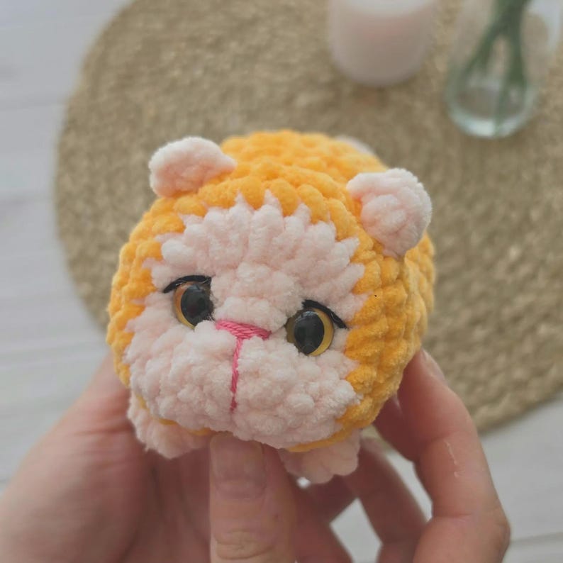 May include: A small, round, crocheted plush toy shaped like a cat. The body is made of soft, textured orange yarn, while the face is light pink. It features two large, dark, glossy eyes with amber highlights and a small, stitched pink nose.