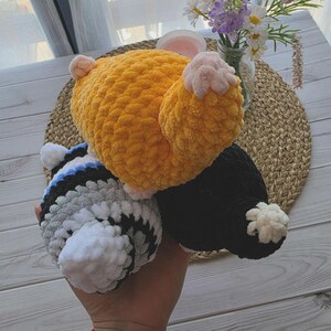 May include: Three plush crochet toys made of soft yarn. One toy is bright orange with light pink accents, one is black with a small white tail, and one features horizontal stripes in white, gray, and black with a white tail. All have a textured finish.