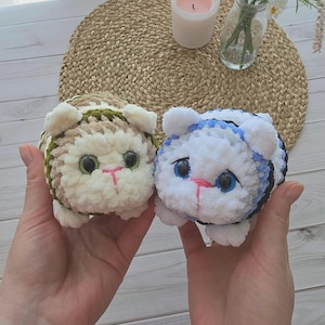 May include: Two round, plush, crocheted cat toys. The left cat is beige and white with green eyes and a pink stitched nose. The right cat is white and blue with black stripes, blue eyes, and a pink stitched nose. Both have small ears and paws.