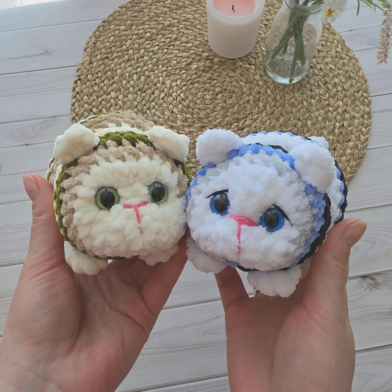 May include: Two round, plush, crocheted cat toys. The left cat is beige and white with green eyes and a pink stitched nose. The right cat is white and blue with black stripes, blue eyes, and a pink stitched nose. Both have small ears and paws.