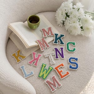 Pode incluir: Uma coleção de patches de letras de feltro coloridos em vários tons, incluindo rosa, azul-petróleo, verde, amarelo, roxo, laranja e azul. Cada letra apresenta uma borda branca e uma superfície texturizada, dispostas ao lado de um livro aberto.