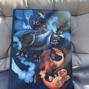 Pode incluir: Um fichário de cartas colecionáveis VaultX azul-escuro com uma grande criatura preta semelhante a um dragão com chamas azuis e duas criaturas menores, cor de laranja, semelhantes a lagartos com caudas de fogo na capa frontal.