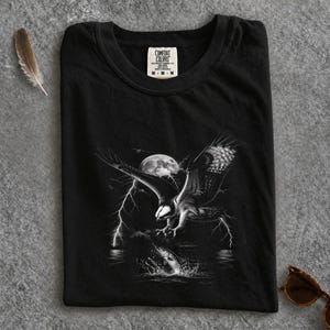 Puede incluir: Una camiseta negra con un estampado gráfico de un águila en vuelo sobre un pez saltando del agua. La escena incluye una luna llena y rayos en el fondo. La etiqueta interior del cuello muestra el texto Comfort Colors M M M.