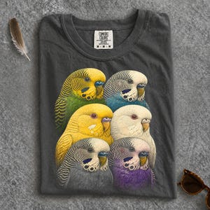 May include: A charcoal gray t-shirt featuring a graphic print of six budgerigar parakeets in various colors including green, yellow, blue, white, and purple. The shirt has a Comfort Colors brand label at the inner neckline and short sleeves.