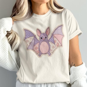 May include: A cream-colored short-sleeve t-shirt featuring a central graphic of a bat designed with a patchwork aesthetic. The bat illustration incorporates various pastel purple and pink patterns, including gingham, polka dots, and floral prints.