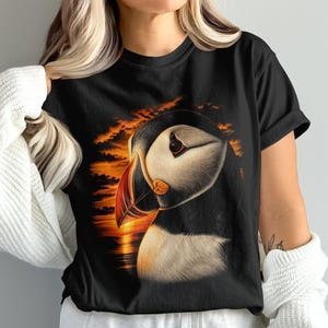 Może przedstawiać: Czarny t-shirt z krótkim rękawem z nadrukiem graficznym przedstawiającym maskonura z kolorowym dziobem. Ptak znajduje się na tle żywego, pomarańczowo-żółtego zachodu słońca z chmurami i linią horyzontu, co tworzy kontrastowy i wyrazisty wzór.