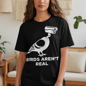 Black t-shirt with white graphic of a bird with a security camera for a head, text reads "BIRDS AREN'T REAL".