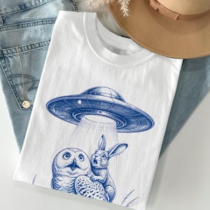 May include: A white t-shirt featuring a blue graphic print of a flying saucer beaming light down onto a snowy owl and a rabbit. The shirt is layered with light blue denim jeans, a tan wide-brimmed hat, and a delicate gold chain necklace.