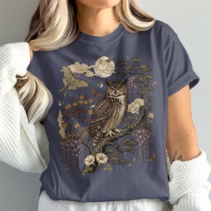 Op de afbeelding: Een donkergrijs t-shirt met een gedetailleerde grafische print van een bruine uil op een boomtak. Het ontwerp bevat een grote nachtvlinder, een volle maan, wolken en hangende bloemenelementen in beige, bruine en gedempte paarse tinten.