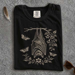 May include: A black t-shirt featuring a detailed, light-coloured graphic of a bat hanging upside down from a branch. The design includes three moths and floral elements surrounding the bat. The shirt has a Comfort Colors brand label at the neck.
