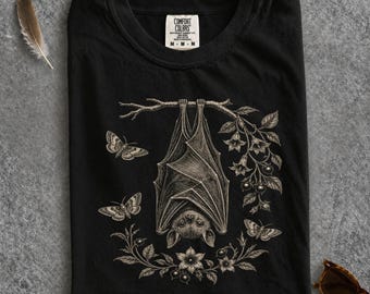 Peut inclure: Un t-shirt noir avec un motif détaillé de couleur claire représentant une chauve-souris suspendue à une branche. Le dessin comprend trois papillons de nuit et des éléments floraux entourant la chauve-souris. Étiquette Comfort Colors au col.