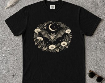 May include: A black short-sleeve t-shirt featuring a central graphic of a bat with outstretched wings beneath a crescent moon, surrounded by a circular wreath of white morning glory flowers and fern leaves. The shirt has a Comfort Colors label.
