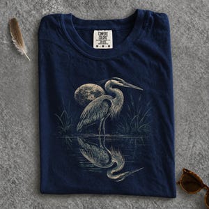 Könnte beinhalten: Ein marineblaues T-Shirt mit einer grafischen Illustration eines Reihers, der im Wasser steht, mit einem Vollmond im Hintergrund. Der Vogel spiegelt sich im Wasser darunter. Das T-Shirt hat ein Comfort Colors Markenetikett am Innenkragen.