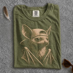 May include: An olive green t-shirt featuring a light brown graphic illustration of a bat wearing a baseball cap. The shirt has a Comfort Colors brand label at the inner neckline. The design shows the bat from the chest up with wings visible.