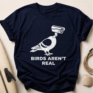 Navy t-shirt with a graphic of a pigeon wearing a surveillance camera for a head, text reads "BIRDS AREN'T REAL".