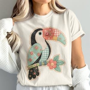 Può includere: Una t-shirt color crema con una grande grafica centrale di un tucano disegnato con un'estetica patchwork. L'uccello è composto da vari motivi di tessuto, tra cui stampe floreali, quadretti vichy e pois in tonalità verde acqua, rosa e nero, appollaiato su un ramo con un fiore.