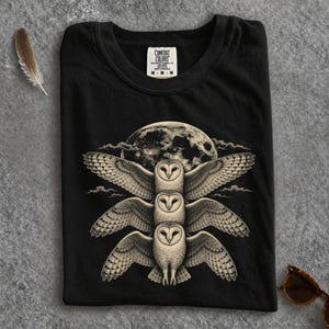 Könnte beinhalten: Ein schwarzes T-Shirt mit einem Grafikdruck von drei gestapelten Schleiereulen mit ausgebreiteten Flügeln vor einem großen Vollmond und Wolken. Das T-Shirt hat ein weißes Nackenetikett mit dem Text Comfort Colors, M und weiteren Details.