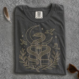 May include: A charcoal grey t-shirt featuring a gold-toned line art graphic of a snake coiled around binoculars, surrounded by birds, stars, and leafy branches. The inner neck label displays the text Comfort Colors, 100% cotton, and size M.