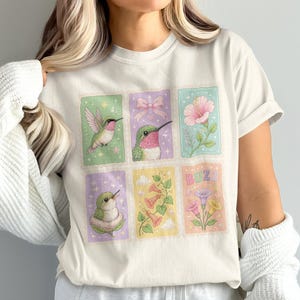 Peut inclure: Un t-shirt couleur crème arborant une impression graphique composée de six panneaux rectangulaires. Les panneaux présentent des illustrations de colibris, un nœud rose, une fleur d'hibiscus, des fleurs en trompette et le mot BUZZ en couleurs pastel.