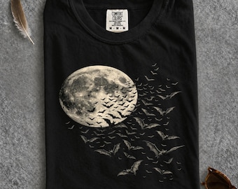 Peut inclure: Un t-shirt noir avec une impression graphique représentant une grande pleine lune détaillée sur le côté gauche, accompagnée d'un essaim de nombreuses petites chauves-souris volant à travers la lune vers la droite. L'étiquette indique Comfort Colors.
