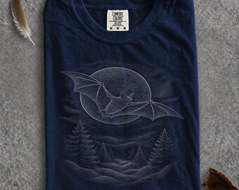 May include: A navy blue t-shirt featuring a white line art graphic of a bat flying in front of a full moon above a forest of pine trees. The shirt has a crew neckline and a Comfort Colors brand label printed on the inside of the collar.
