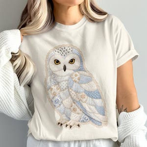 Può includere: Una t-shirt a maniche corte color crema con una grande grafica di un gufo delle nevi. Il gufo è composto da vari tasselli a motivi blu chiaro, bianchi e beige che ricordano un design patchwork, con occhi giallo brillante e artigli scuri.