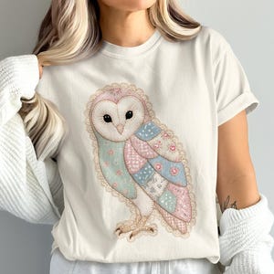 May include: A cream-coloured short-sleeve t-shirt featuring a central graphic of a barn owl. The owl is designed with a patchwork pattern in soft shades of pink, blue, and white, accented with delicate lace-like trim along its outer edges.