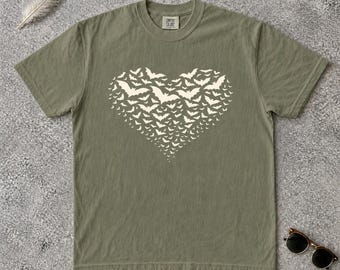 May include: An olive green short-sleeve t-shirt featuring a large, cream-colored graphic of a heart shape composed of numerous small, flying bat silhouettes. The shirt has a crew neckline and a Comfort Colors brand label printed on the inner collar.
